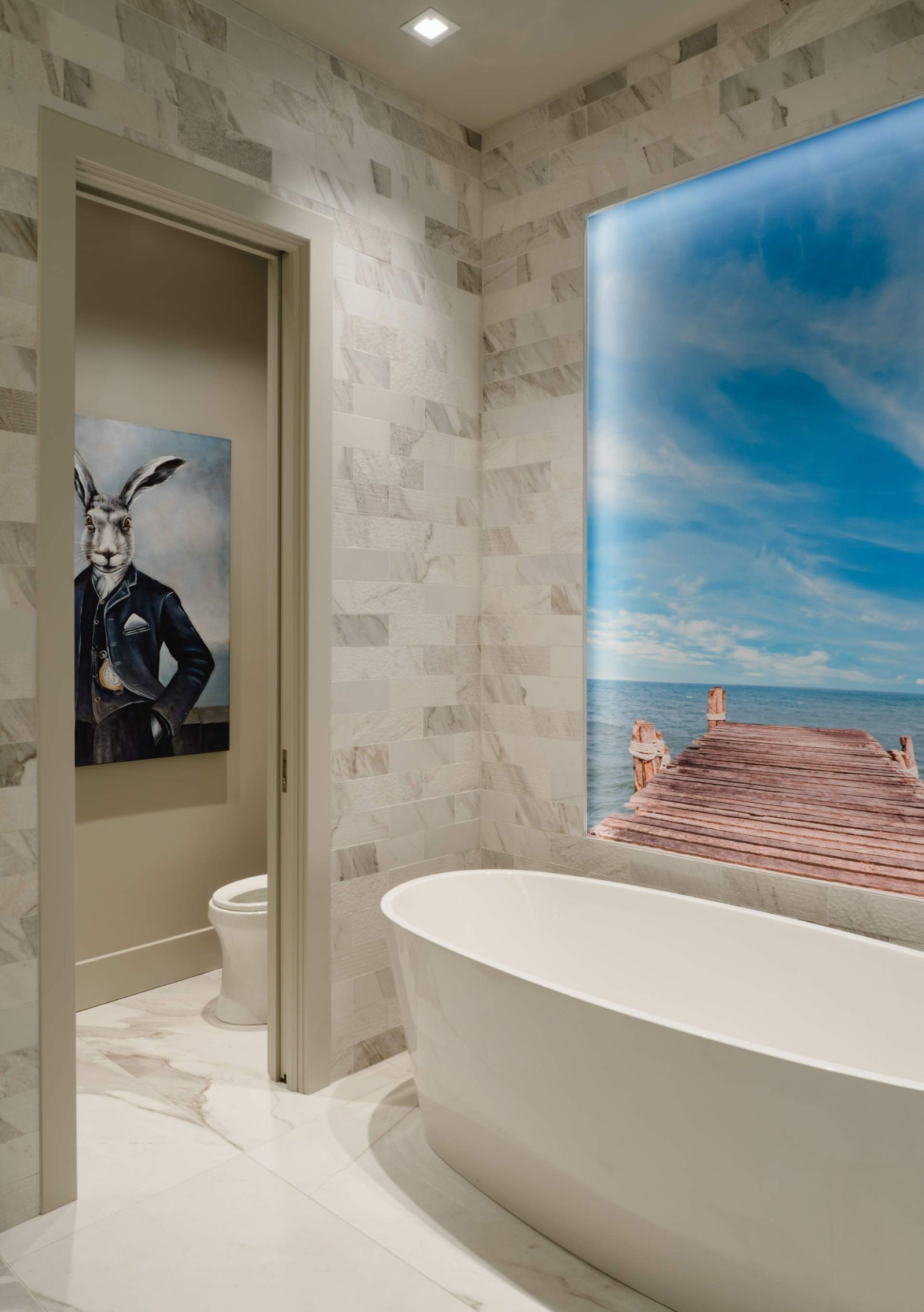 Freestanding tub beneath a backlit ocean mural in a marble bath