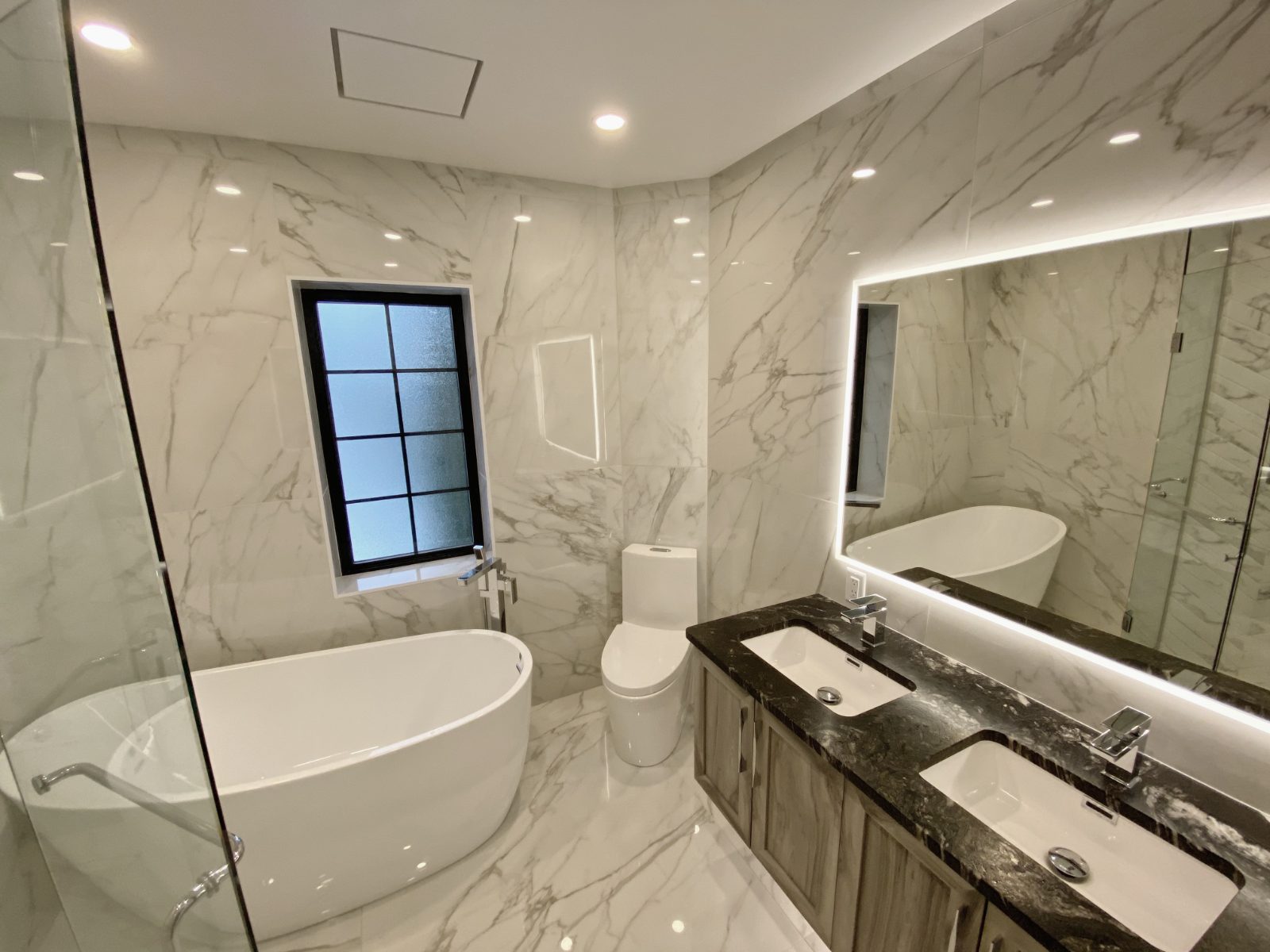 Bath wrapped floor to ceiling in marble with freestanding tub and backlit mirror